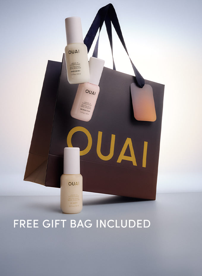 Three OUAI Leave in conditioner bottles arranged around a large dark brown shopping bag with gold OUAI lettering and a gradient hangtag, on a light gray studio background. White text at the bottom reads 'FREE GIFT BAG INCLUDED'.