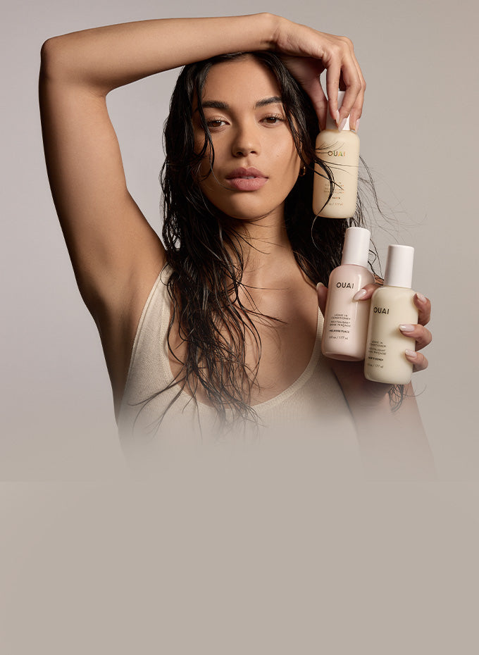 A woman with long dark hair holds three O'NAI hair care bottles against a neutral beige studio background.