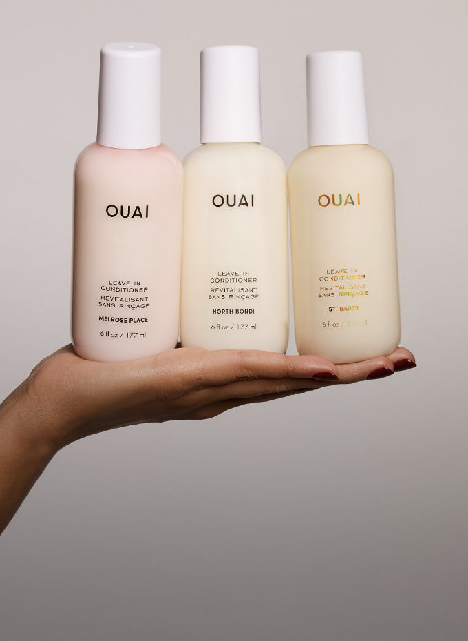 Three OUAI leave-in conditioner bottles in soft pastel pink, cream, and light beige, held in a person's hand against a neutral gray background.