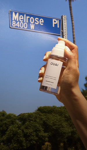 Hand holding a OUAI product under a Melrose Place street sign with trees in the background