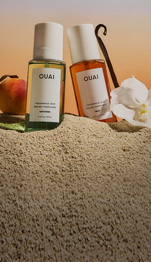 Two OUAI perfume bottles on sand with a peach and orchid in the background